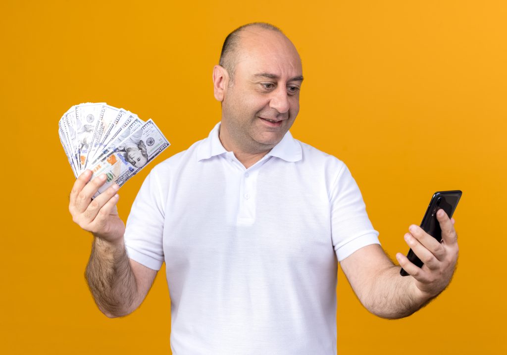 Smiling Casual Mature Man Holding Cash Looking Phone His Hand Isolated Yellow Wall Scaled